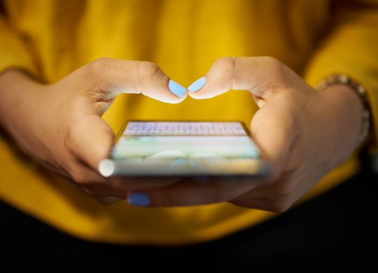 Young woman using cell phone to send text message on social network at night. Closeup of hands with computer laptop in background