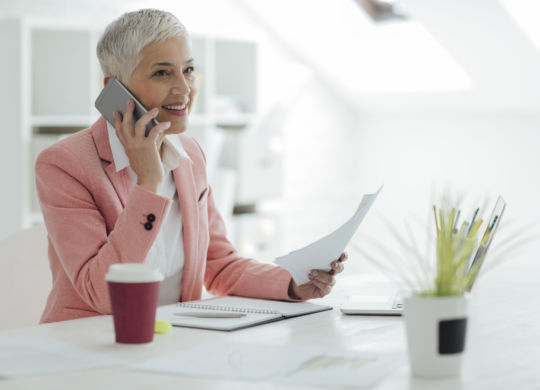 Mature businesswoman working in her new modern office. Sitting by the desk and talking on the phone with client. She is happy and cheerful. Holding papers in the hand.